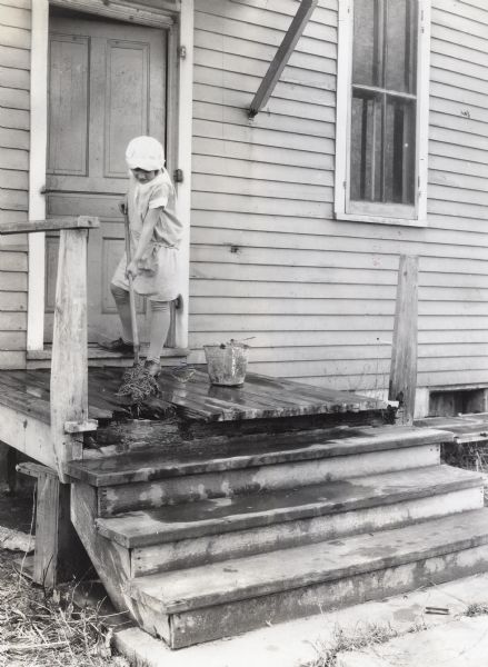A woman wearing a dress, hat, and stockings uses a mop and bucket to wash a damaged wooden porch attached to a farmhouse.