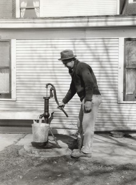 A man uses a well to pump water into a metal bucket while standing in front of a farmhouse.