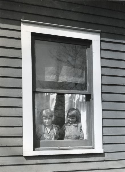 Exterior view of two girls standing at the closed window of a farmhouse while their parents are away. Photograph taken for International Harvester's Agricultural Extension Department to illustrate various farm hazards.