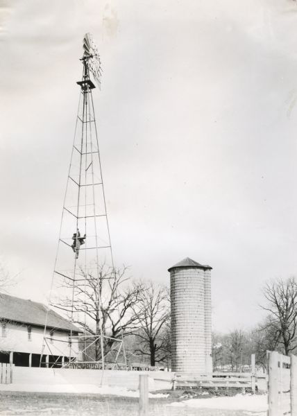 A man climbs the base of a windmill at Cutten Farms. Fences, a farm building, and a silo are around the windmill.