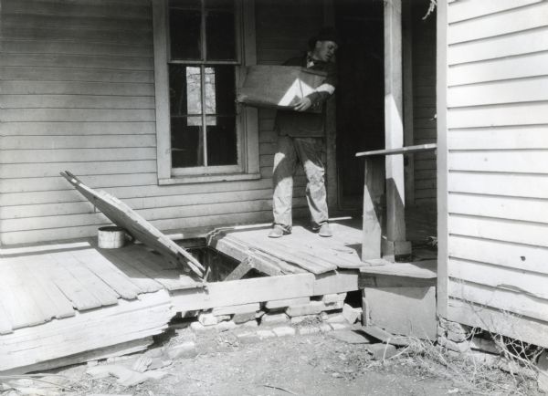 A man holding a large crate in his arms approaches an open bulkhead cellar door while walking on the porch of a farmhouse.