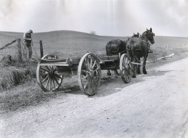 A wagon hitched to two horses is standing alongside a dirt road as a man uses a hoe or shovel in the field to the left.