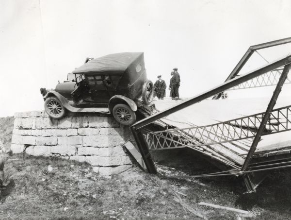 Men examine the wreckage of an automobile resting on the edge of a foundation near a collapsed bridge. A portion of a crane can be seen in the background attached to the automobile.