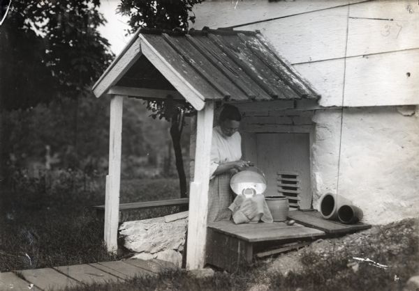 A woman standing under a rooftop near a doorway pouring liquid through what appears to be a cheesecloth strainer and into a clay pot.