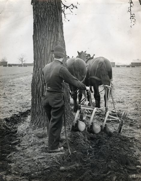 Rear view of a man using an Oliver plow pulled by a team of two horses to work around a tree in a field. A railroad train is in the background.