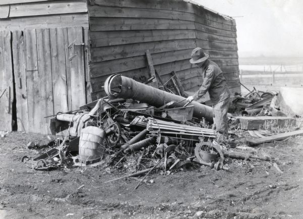 A man is looking through a scrap heap full of metal and wooden parts piled beside a shed or barn.