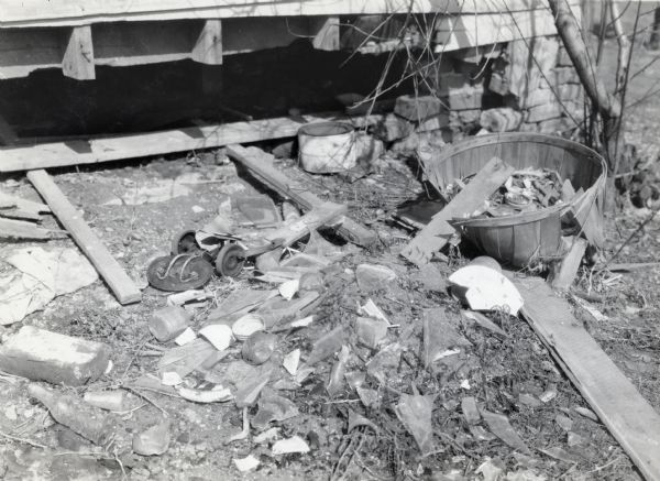 A refuse pile consisting of glass, a broken basket, bottles, cans, and other items behind a farmhouse. A crumbling foundation is in the background.