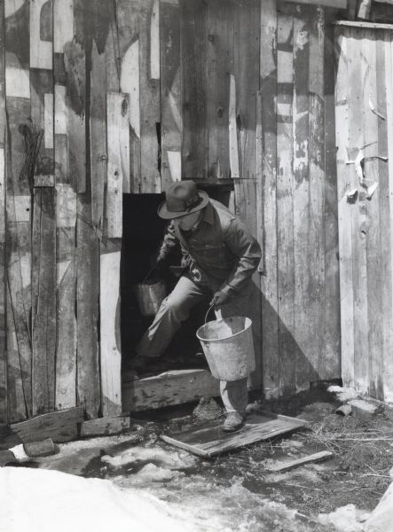 A man holding a metal pail in each hand ducks as he is exiting a low barn door. Snow and ice are just outside the door.