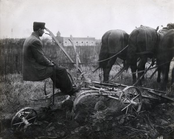 Side view of a man driving a team of three horses pulling an Oliver plow across a field. A large building, houses, and a water tower are in the background.