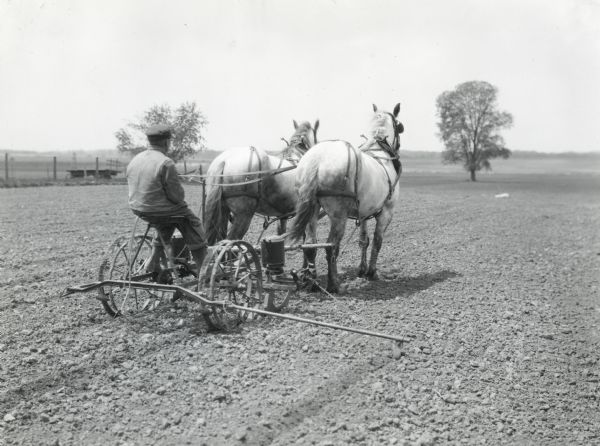 A man is using a planter drawn by a team of two horses to work in a field at Bainridge Farm. A fence and what appears to be a wagon bed are in the background.