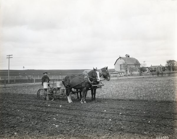 A man is operating a horse-drawn McCormick corn planter with a fertilizer attachment in a field. A dirt road, barn and farmhouse are in the background across a road.