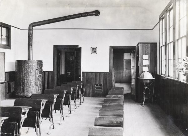 Interior view of empty classroom of rural school. On the far left is a large, round, wood stove with decorative metal work.