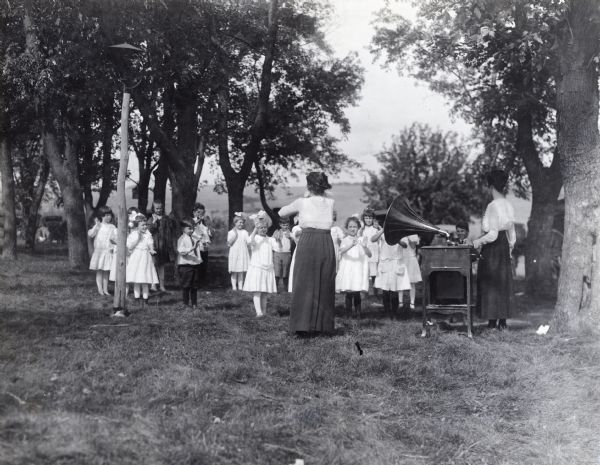 Miss Streeter leading school children in the "shoemaker's song". Another woman is standing near a Victrola. The children are outdoors under trees. Possibly in a rural schoolyard.