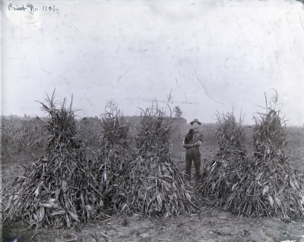 Farmer standing outdoors among corn shocks. He is holding a small dog in his arms, and is wearing work gloves and a hat.
