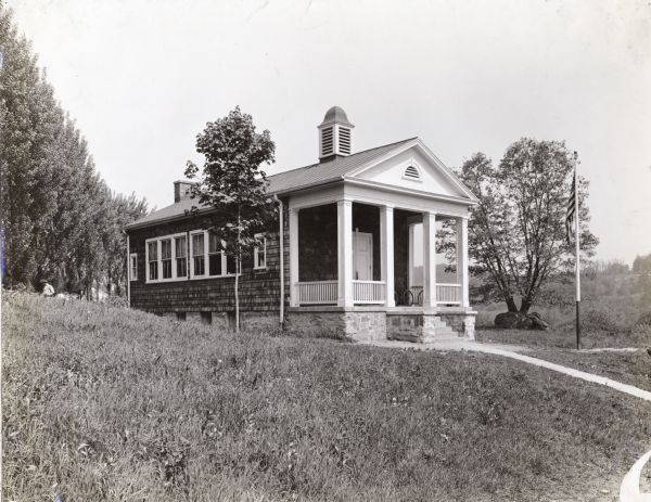 Exterior photograph of schoolhouse. Original caption reads: "one room school cost $3786." The building has a stone foundation, a roof vent, and a chimney at the back. The front porch has columns and railings, and there is a gable vent above the porch on the front of the building. Two bicycles are parked on the porch. A flag pole is on the right, and a person wearing a hat is standing in the background on the left side of the school.