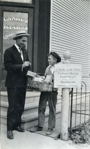 Young Charles Vosmik posing outside a candy and ice cream shop with a basket of goods to sell to a man standing on a sidewalk. The man is holding a coin in his hand to pay the boy. There is a sign on a post that reads: "Cook County / School Home Project / Business."