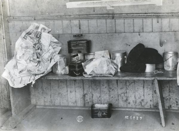 Lunch pails, newspaper-wrapped packages, and hats are stacked on a bench inside Sedan Prairie School. The paper hanging on the wall above the bench reads: "Only Red Cross Members are Permitted to Display This Service Flag."