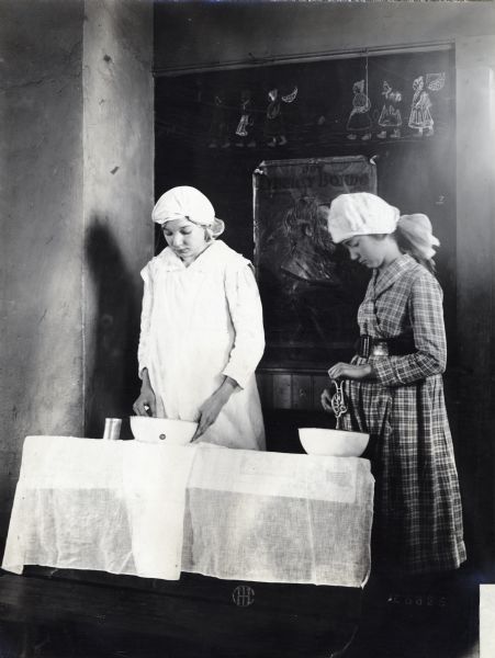 Two girls mixing food in a bowl while preparing lunch at Sedan Prairie School. A poster reading: "Buy Liberty Bonds" is on the blackboard behind them, and what appear to be Dutch figures are drawn in chalk above it.