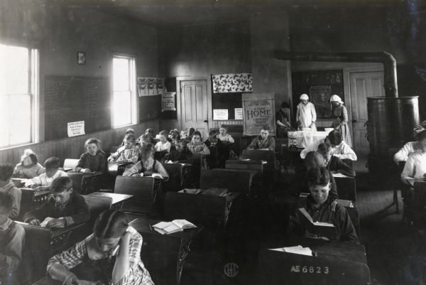 Students sit at desks inside Sedan Prairie School while others stand in the back of the classroom to prepare lunch. The poster hanging on the back wall reads, "Agricultural Lecture Charts. A Good Home Provides Comfort, Profit, Pleasure." A large round wood stove is in the back right corner of the room.