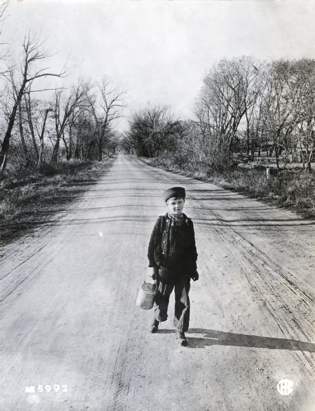 A boy carrying a metal lunch pail walking along a dirt road.