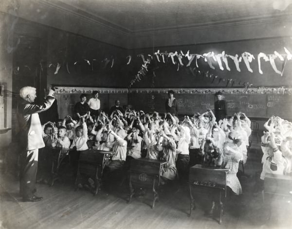 A group of children sitting at desks raising their arms while being led in a classroom activity by a male instructor. Four women and one man are sitting and standing watching in back of the classroom. Chalkboards line the walls of the room and paper decorations are hanging from the ceiling.