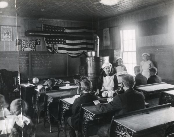 Girls serving trays loaded with mugs of soup to students seated at desks in a classroom. A chalkboard, portraits of presidents, and an American flag are in the background. A large round woodstove is in the back corner of the room.