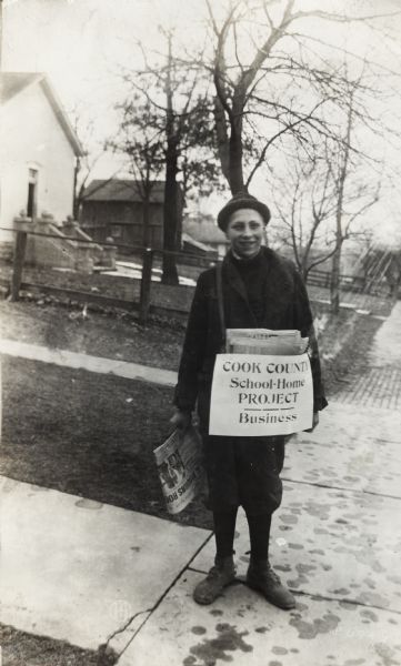 A boy wearing a coat and hat standing on a sidewalk while delivering newspapers to nearby homes. Attached to his newspaper bag is a sign reading: "Cook County School-Home Project - Business."