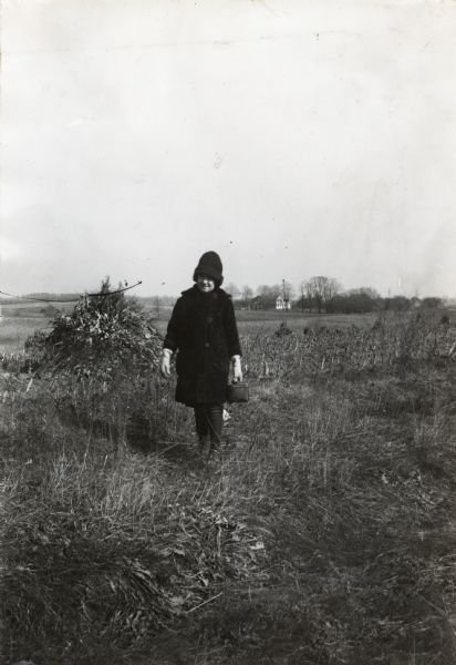A girl wearing a hat, winter coat and gloves walking through a field while carrying a lunch pail.