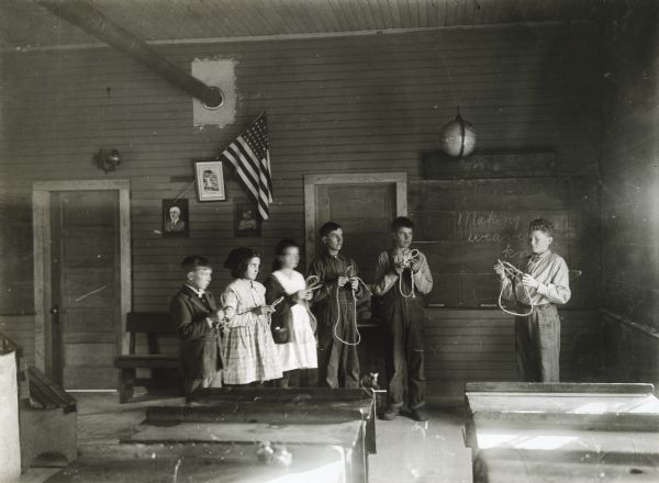 A group of students learning to tie knots in pieces of twine in a classroom. A chalkboard, American flag, and several portraits are hanging on the wall in the background.