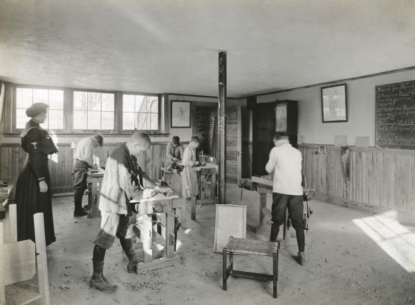 View of a teacher watching over a group of male students as they practice woodwork at benches inside a classroom. The text on the blackboard hanging on the wall reads: "Rules for Planing."