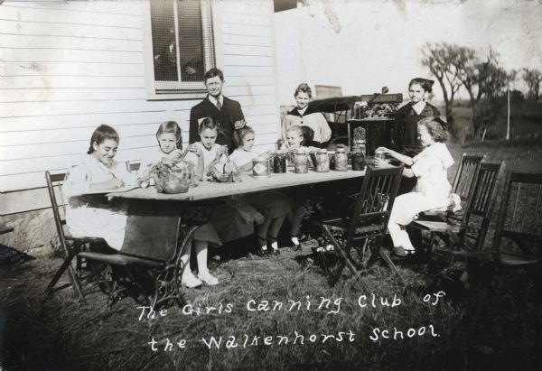 A group of girls from the Canning Club of Walkenhorst School sitting and standing around a table outdoors to display their canned goods. A man wearing eyeglasses, perhaps a teacher, is standing behind them.