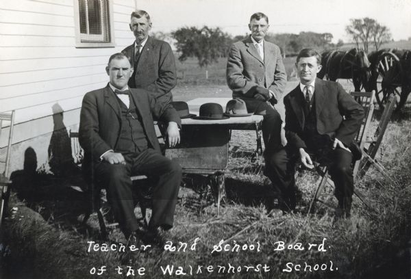 Teacher and school board of the Walkenhorst School sit around a table outdoors near a building. The men have set their hats on the table, and a horse-drawn wagon is in the background.