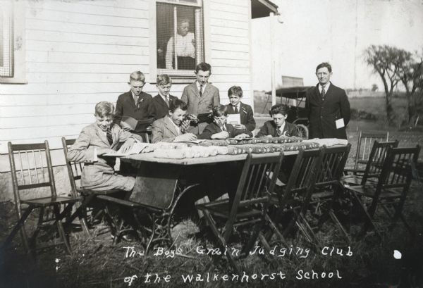 A group of boys from the Boys Grain Judging Club at Walkenhorst School sitting around a table outdoors to look at cobs of corn. A man, wearing eyeglasses, perhaps a teacher, is standing near the table, and a woman is looking out from a window in the building behind the boys.