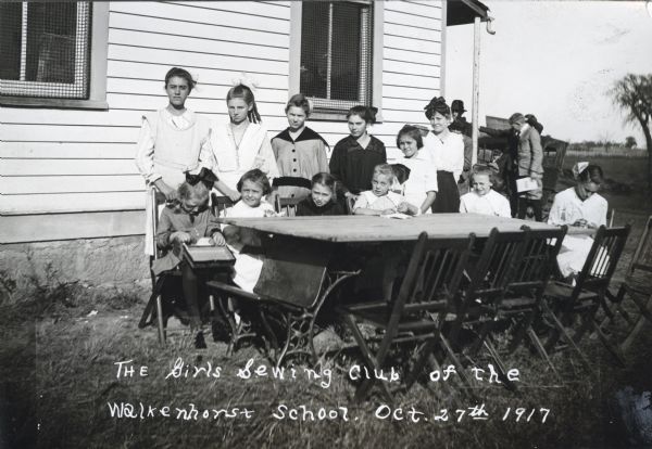 Members of the Girls Sewing Club of Walkenhorst School sitting and standing near a table set up outdoors near what is probably a school building.