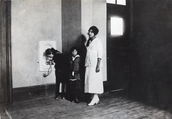 A woman, possibly a school instructor, is standing near two children as they wait in line to use a drinking fountain.