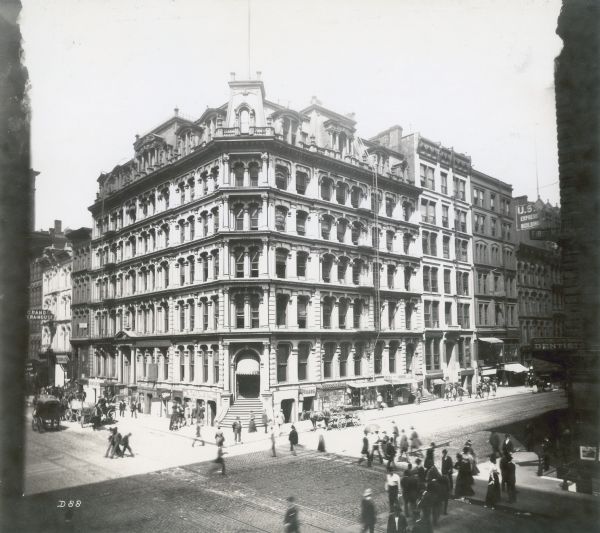 Elevated view of pedestrians and horse-drawn carriages along the streets in front of the city block that housed the McCormick Reaper Building. The building signs read, "Grand Opera House," "Frank's," "W.T. Underwood, Attorney at Law," "The Creditors Agency; National Law & Collection Association," "Cigars," "Shoes - Try a Harkins," and "U.S. Express Building." The sign above the corner door reads, "Reaper Block."