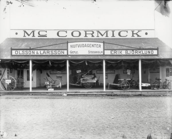 Exterior view of front of a McCormick agricultural machinery dealership in Stockholm, Sweden. Pieces of machinery are arranged on the building's front porch, and bicycles lean against the right wall. Signs on the building read, "McCormick. Olsson & Larsson. Hufvudagenter, Gefle. Stockholm. Erik Bjorklund."