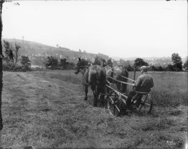 Rear view of a man on a mower driving a team of two horses to work in a field. In the distance are a number of houses on the side of a large hill.