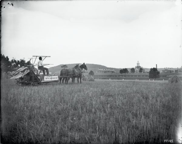 Side view of a man driving a team of horses to pull a McCormick grain binder through a field. In the far background are several large buildings.