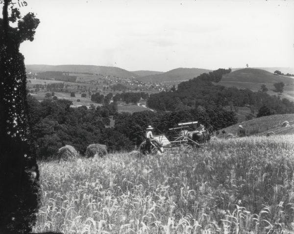 A man is using a horse-drawn McCormick grain binder on a hillside overlooking a valley with houses and other buildings.