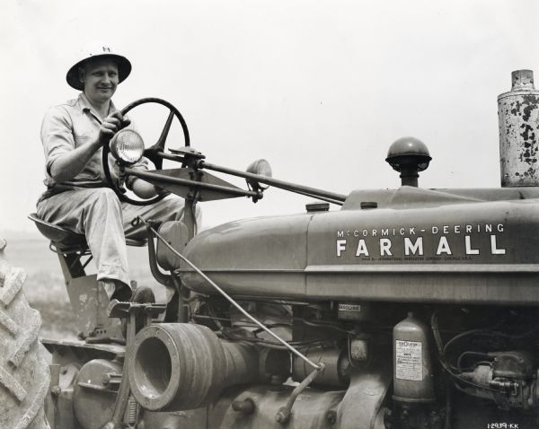 Man wearing an IH pith helmet operating a Farmall M(?) tractor. There are decals on the right side of the tractor.