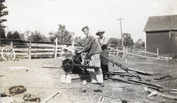 Two men stand in a barnyard assembling what appears to be a piece of agricultural machinery, possibly a grain binder. The men work on top of a wooden crate and use metal parts lying nearby on the ground. A barn and fence are in the background.