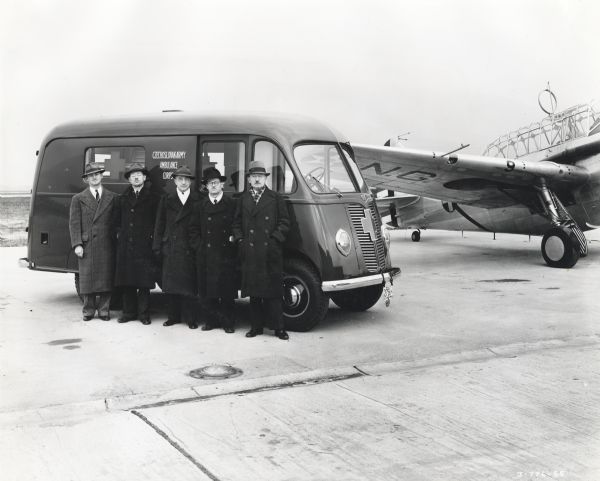 A group of men from the Chicago Czechoslovak Relief Committee are standing against an International D-15-M (Metro) truck sold to Czechoslovakian Relief for service with the Czechoslovakian Army in England. An airplane is in the background on the right.