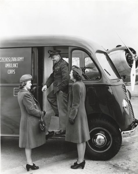 First Lieutenant K.J. Fogle is standing in the cab of an International D-15-M (Metro) truck sold to Czechoslovakian Relief for service with the Czechoslovakian Army in England, while two TWA hostesses are standing outside near the automobile's door. An airplane is parked behind the ambulance.