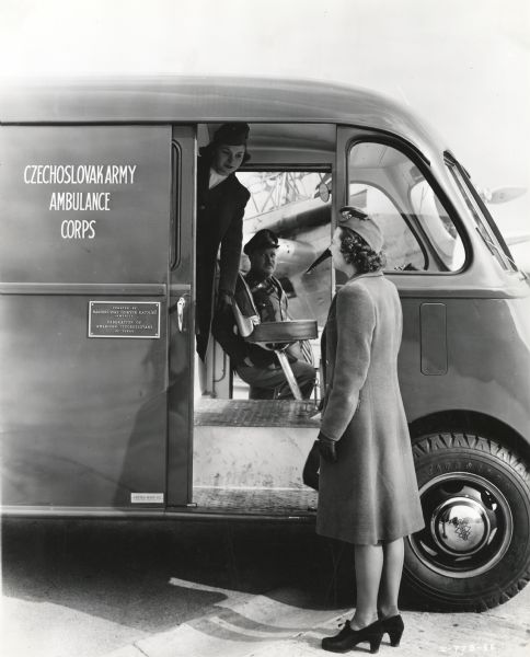 Ruth Ellison, TWA hostess, looks into an an International D-15-M (Metro) truck converted into an ambulance and sold to Czechoslovakian Relief for service with the Czechoslovakian Army in England. Eula Walker, hostess, and First Lieutenant K.J. Fogle, 108th Observation Squadron, look out from inside the vehicle. An airplane is parked in the background.