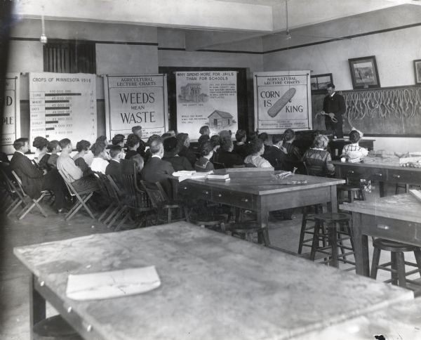 Adults sitting in chairs in a classroom to hear an agricultural extension lecture given by a man standing behind a table or podium at the front of the room. Several posters reading: "Crops of Minnesota 1916," "Weeds Mean Waste," "Why Spend More for Jails Than For Schools?," and "Corn is King" are hanging in the background.