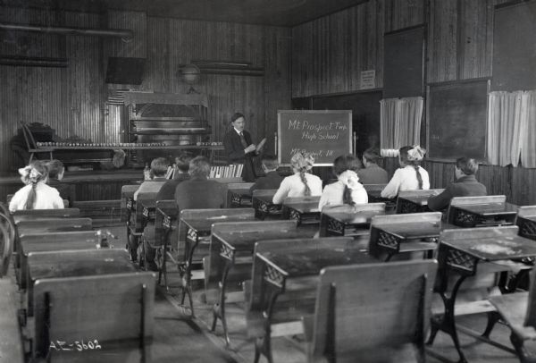 View from the back of a classroom towards a teacher holding an ear of corn while standing near a chalkboard which reads: "Mt. Prospect Twp. High School, Mt. Prospect, Ill." A small group of children are seated at desks near the front of the room.