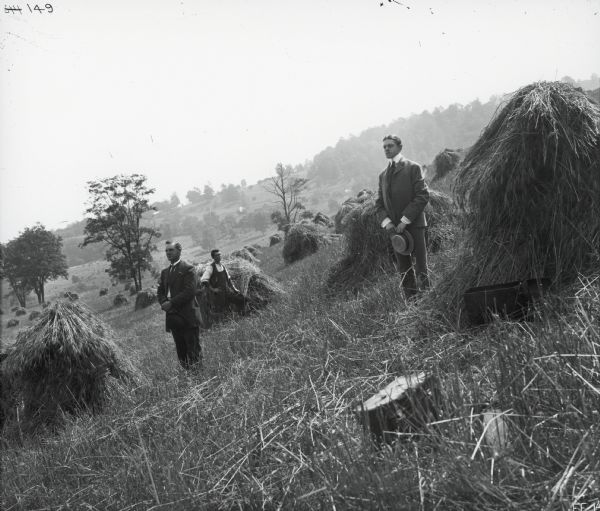 Three men, all looking away from the camera and holding their hats in their hands, are posing while standing in a hillside field next to piles of harvested wheat.