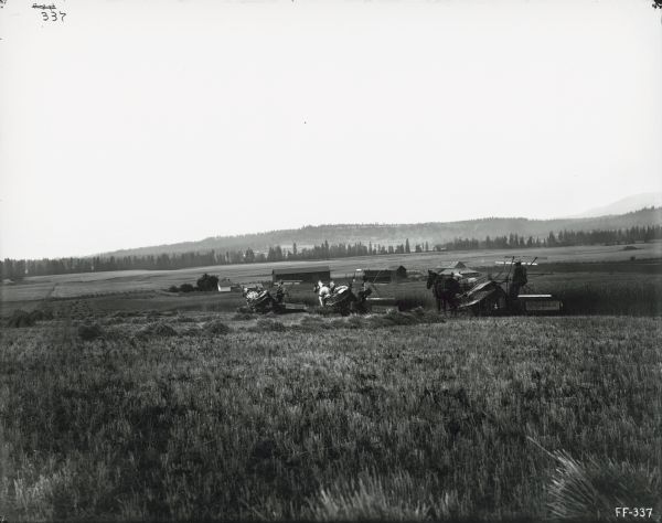 View from a distance of three farmers, and perhaps a child, using horse-drawn McCormick grain binders in a field. Farm buildings are in the middle distance.