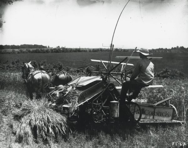 A man wearing a wide-brimmed hat is using a McCormick grain binder pulled by two horses to harvest a field of wheat.